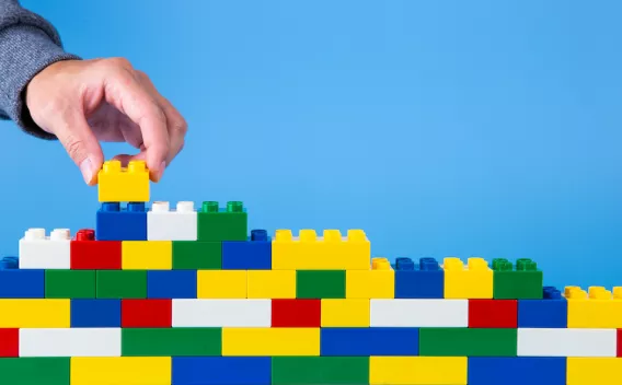 Photo of lego bricks of different colours stacked up with a hand adding another brick, against a blue background.